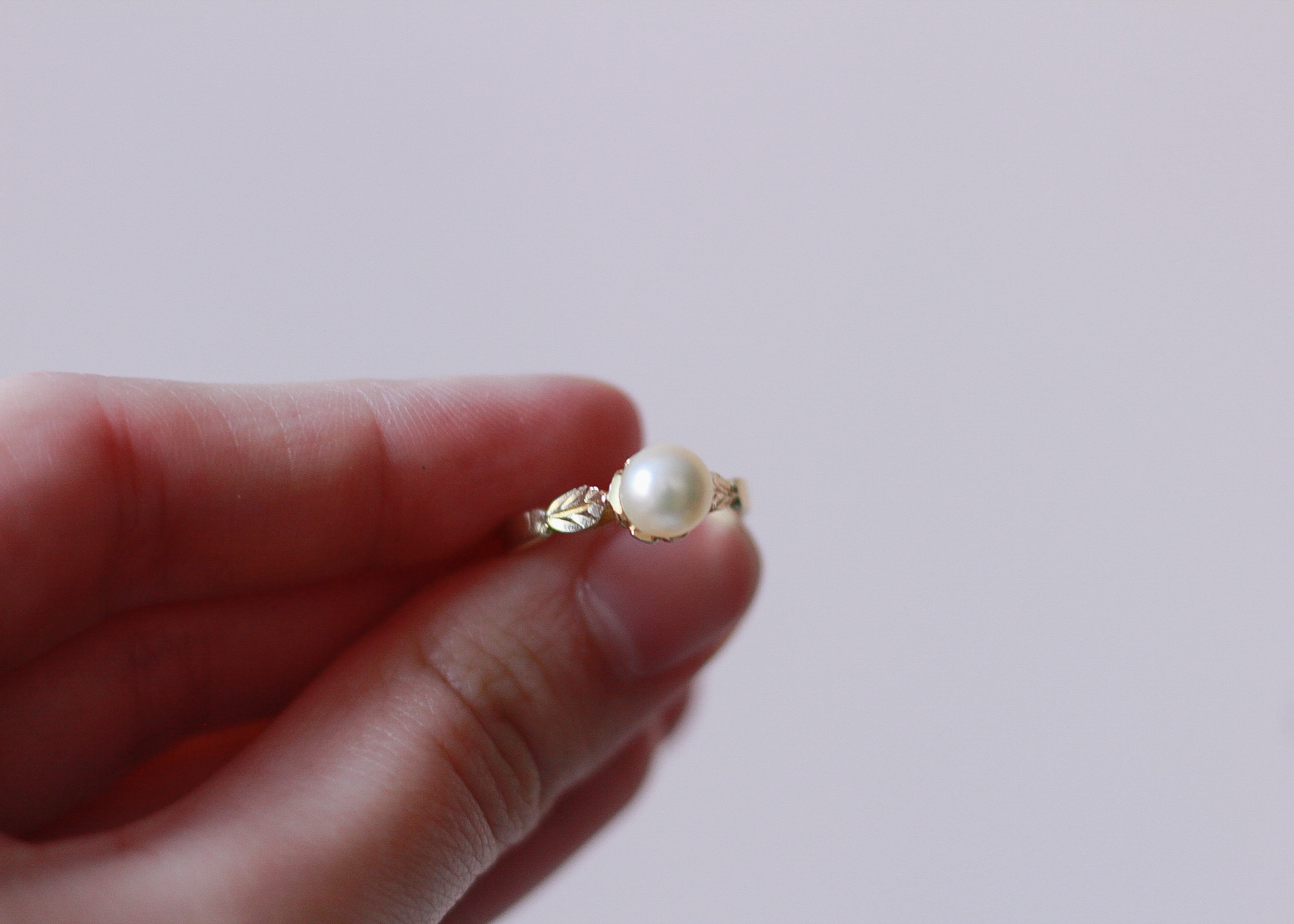 image of a left hand in front of a pink wall, holding a gold ring with a round white pearl on it, as the pearl is facing the camera.